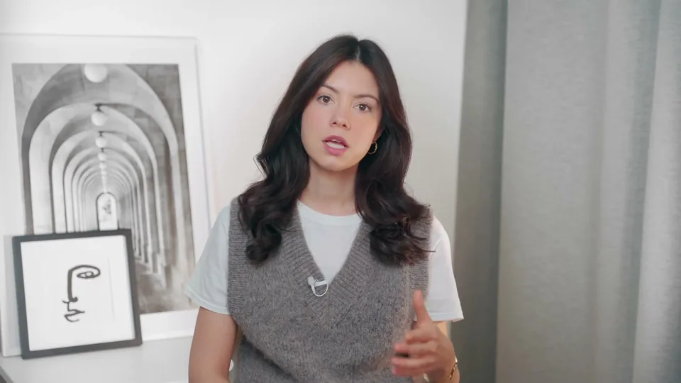 A creator in a grey knit vest talking to the camera in front of a framed print