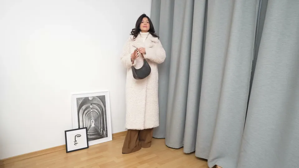 A creator in a long cream coat with a grey bag, full length, by a grey curtain