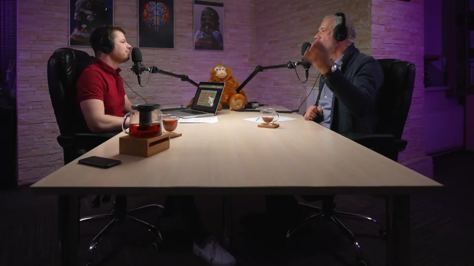 The wide camera: the host and the guest at a long table under purple light, with microphones on arms