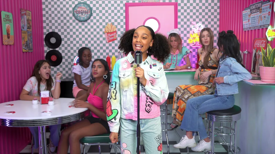 The wide shot: the young singer at a vintage microphone in a pink diner, with kids at a table and at the counter behind her