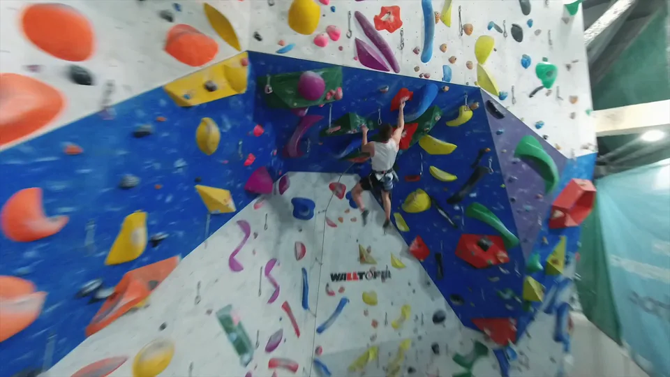 An FPV drone shot along a colorful climbing wall, reaching the climber on an overhang